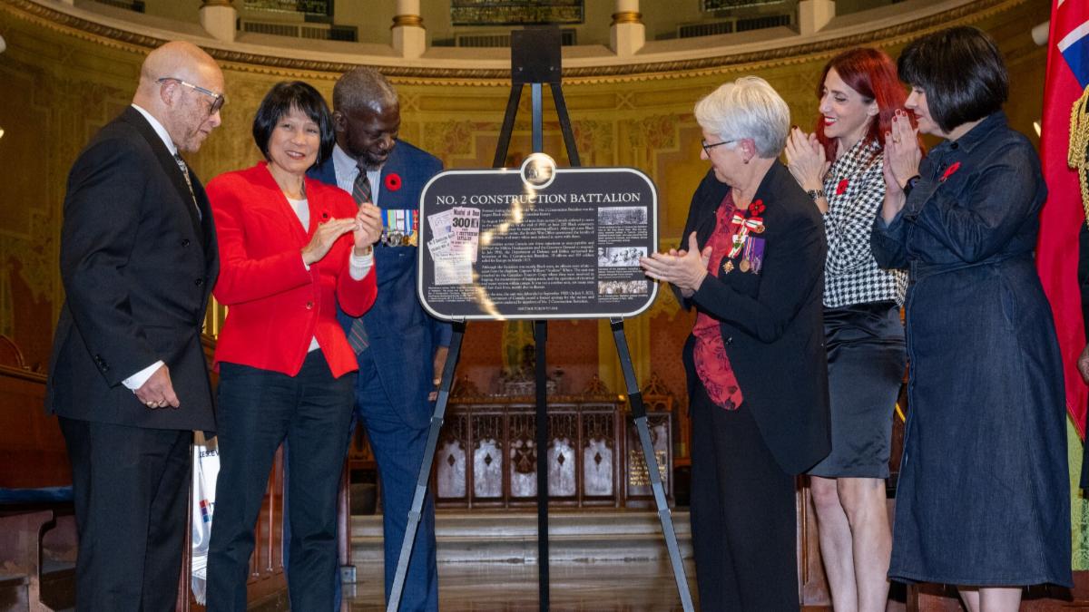 A group of dignitaries and politicians stand and applaud beside a commemorative plaque on an easel.