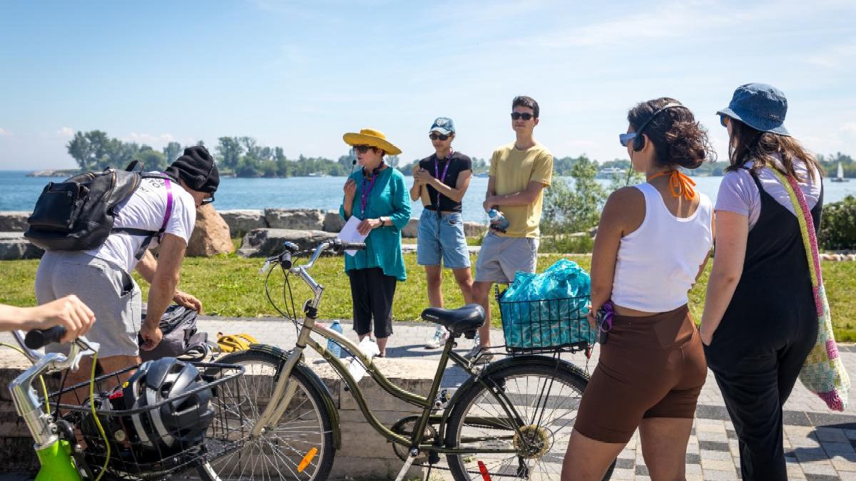 While a lady speaks into a microphone 4 bystanders listen intently while holding on to their bikes.