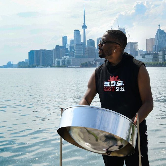 A man stands behind a steelpan. He wears a black tank top. Behind him is a large body of water and the city skyline of Toronto.