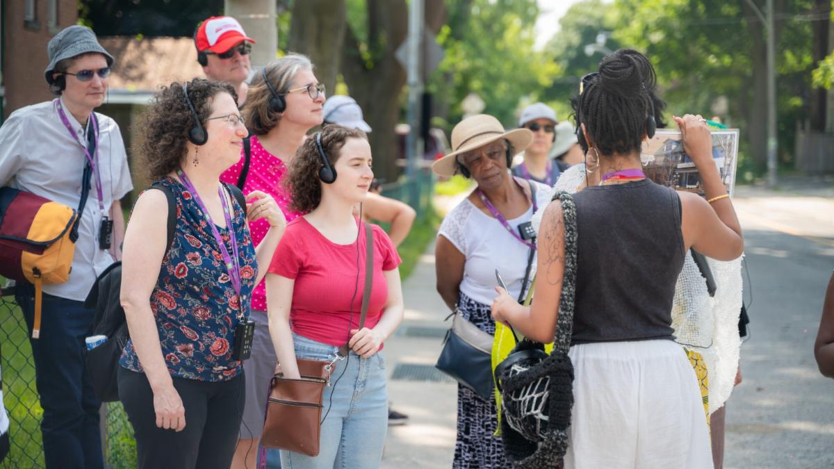 A group of people stand attentively as a guide presents information using a visual aid. The scene takes place in a residential area with trees and houses in the background.