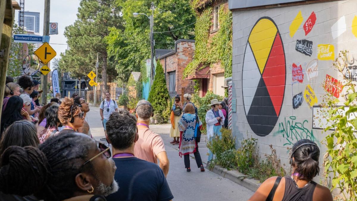 A group of people are standing in a laneway. On the wall of the building to the right of the laneway is a mural of an Indigenous medicine wheel (a circle with yellow, white, red, and black wedges). Large green trees are in the background.