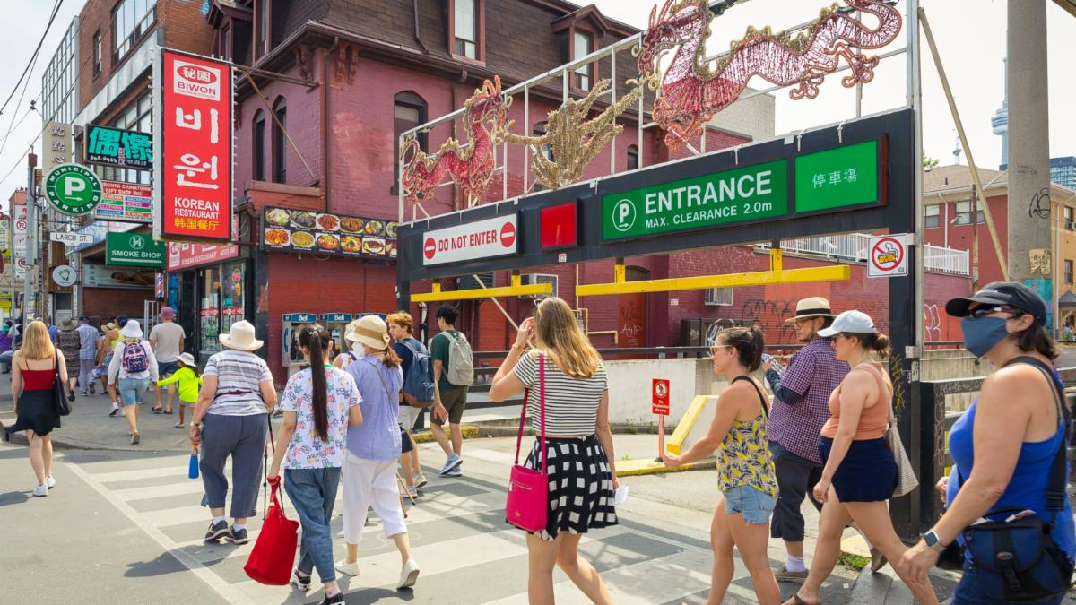 Line of people walk across a city street front. Signage features Chinese characters and parking lot entrance lighting in the shape of two large dragons.