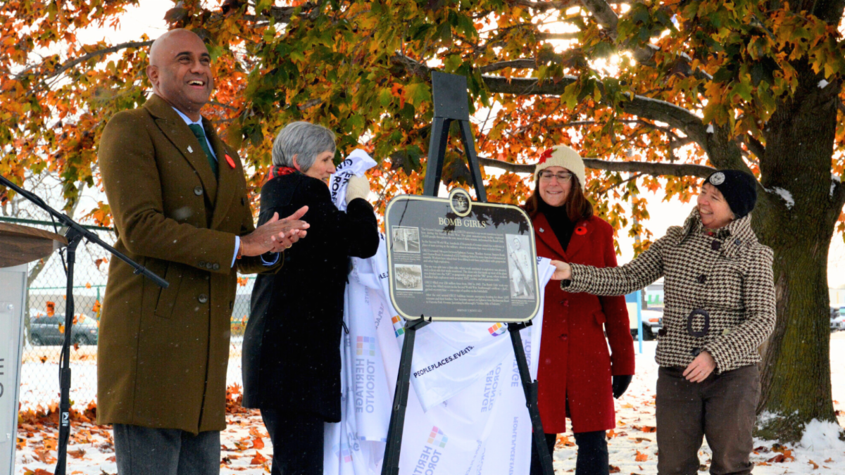 Four people stand around a plaque while the second person from the left removed a white tarp with Heritage Toronto logos from the plaque. The person on the far left is clapping and everyone depicted is smiling.