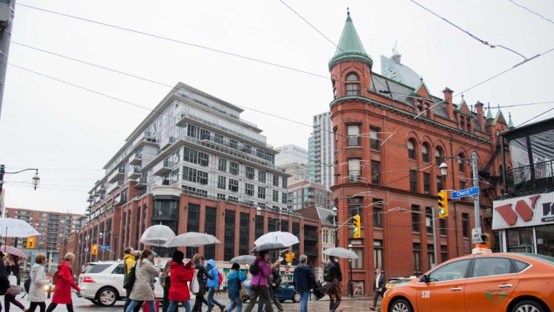 A group of people holding umbrellas cross the road on a busy intersection on a rainy day. The Flat Iron building can features prominently with its distinctive triangular shape.