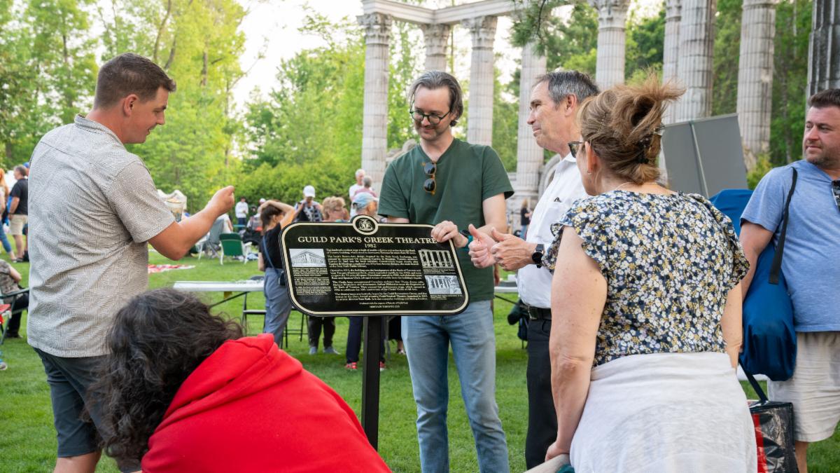 A group of people stand around a plaque displayed on a podium in a park setting. 