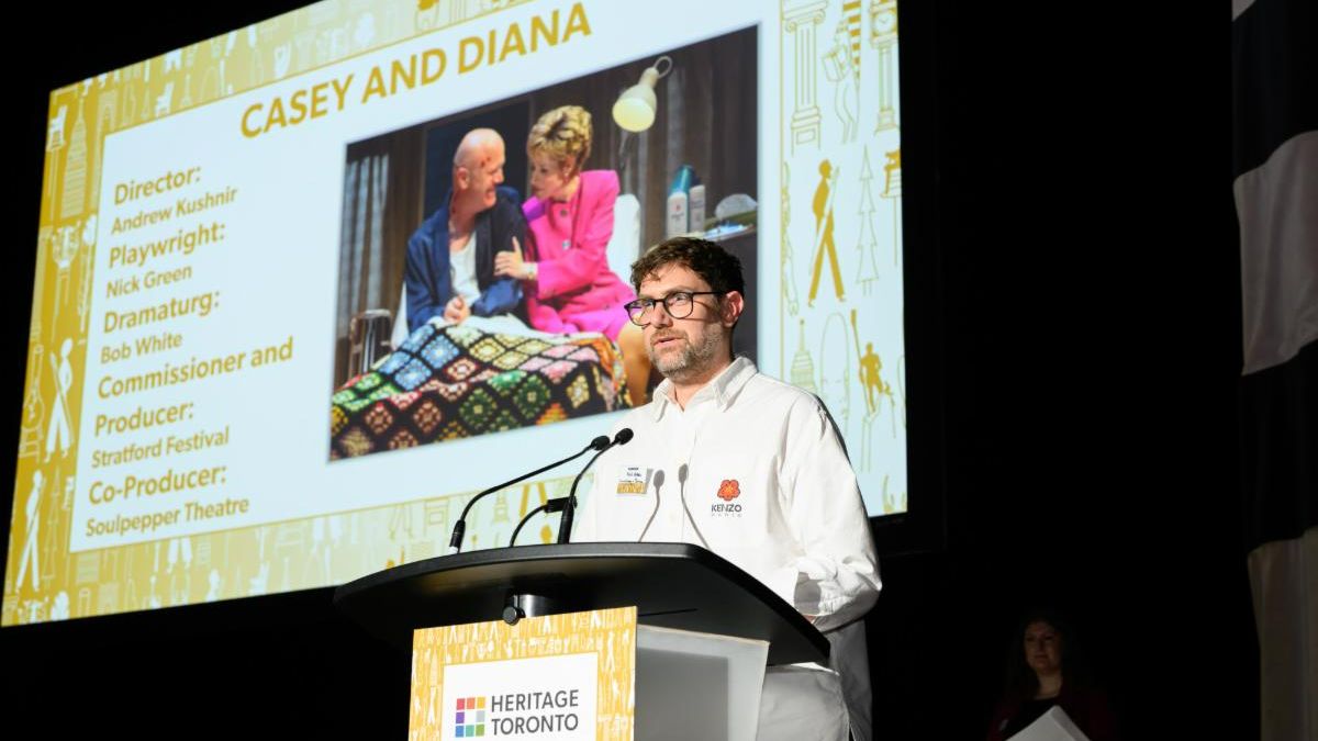 Person at podium speaks into a microphone. Screen behind shows an image with text reading Casey and Diana and names of project team members.
