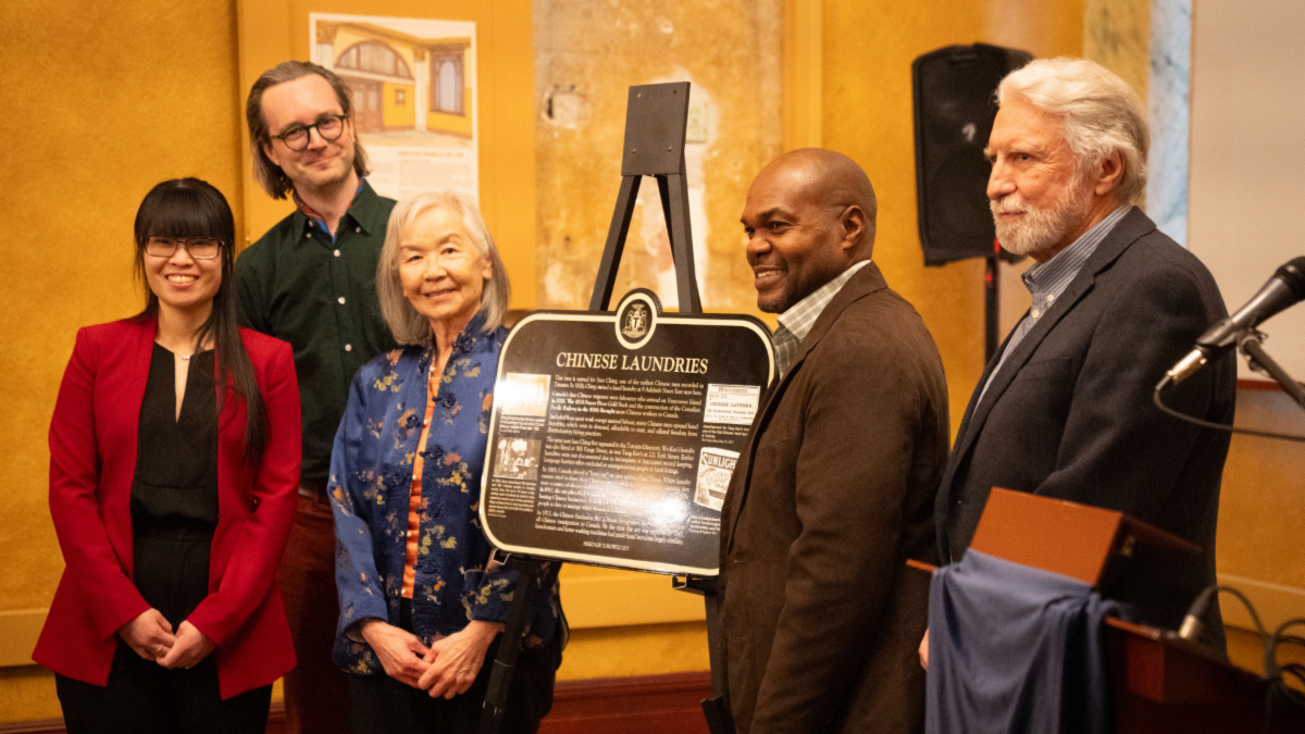 Five people pose with a black and gold plaque in the middle, smiling to the camera.
