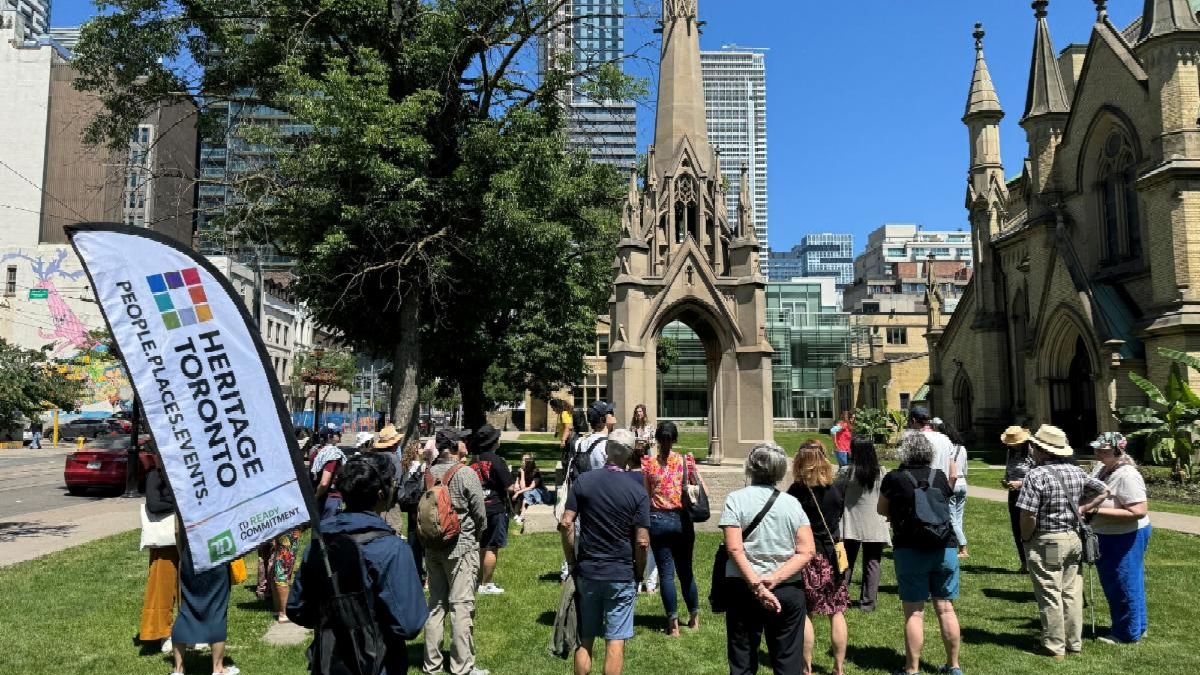 A large group of tour participants gather on the lawn in front a Gothic Revival cathedral on the right. The Heritage Toronto flag is prominent at the left side of the group.