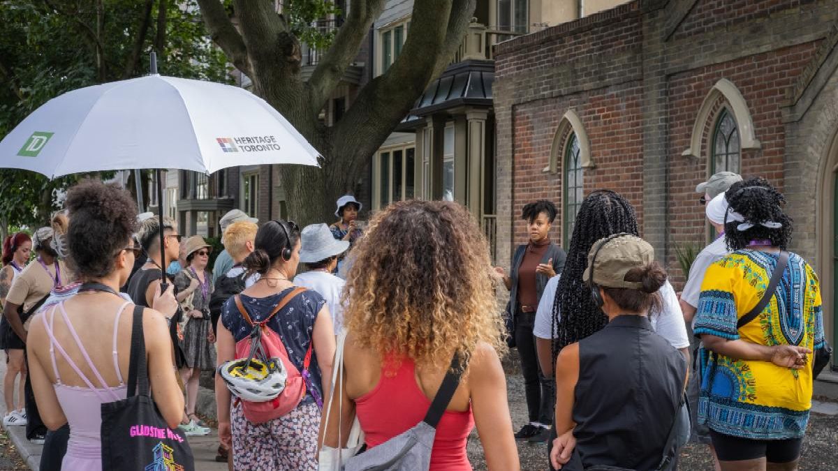 Group of people face away from the camera listening to a woman in the background.