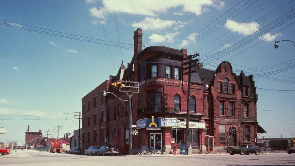 Red brick building with a sign reading Canary Restaurant visible on the front. The building is located at the corner of an intersection where several cars are seen driving or parked along the curbs. 