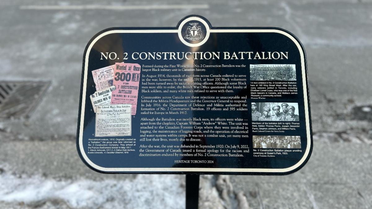 Photograph of a historical plaque titled No. 2 Construction Battalion that is standing on a metal post near the curb of a sidewalk â some snow is on ground near the base of the post.