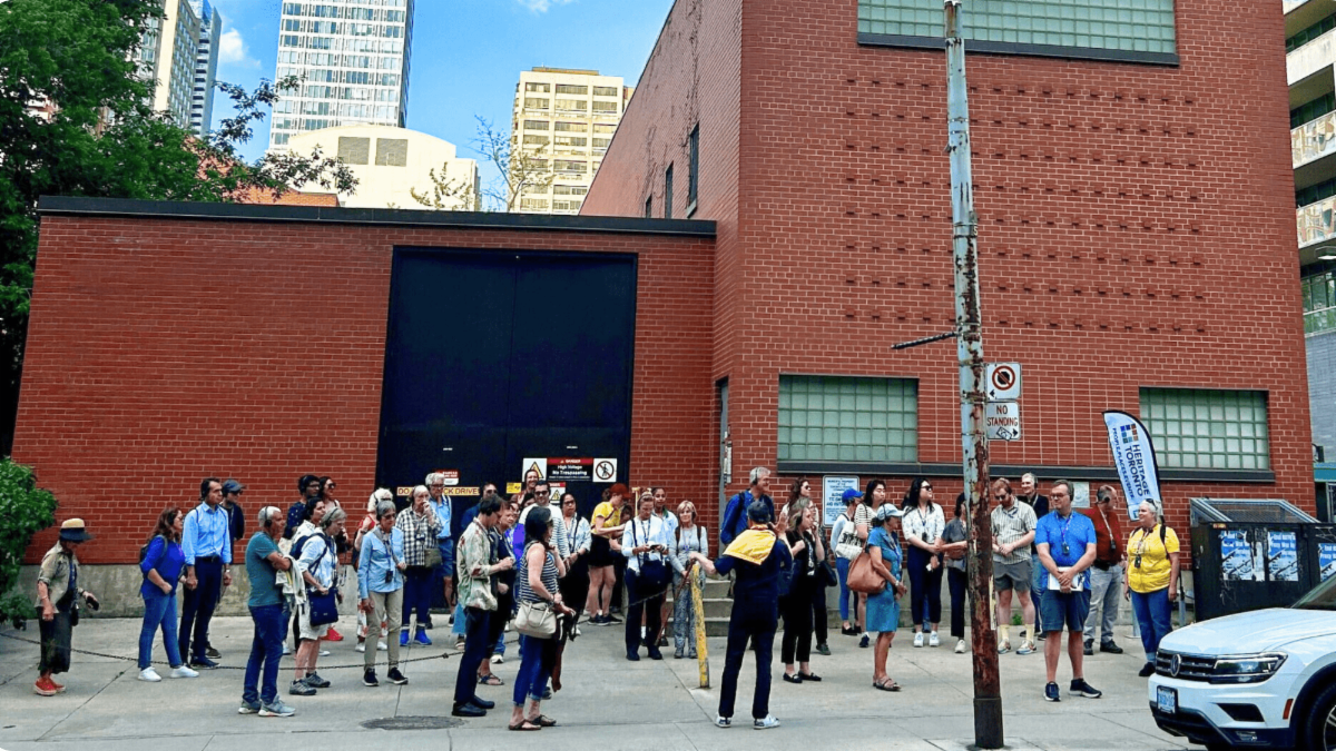 A large tour group is gathered on the sidewalk in front of a brown building on the U of T campus. The left side of the building is a single story with a large black door. The right side of the building is two stories with two small rectangular windows on the lower story and one long rectangular window at the top of the building. The Heritage Toronto flag is located on the right side of the tour group.