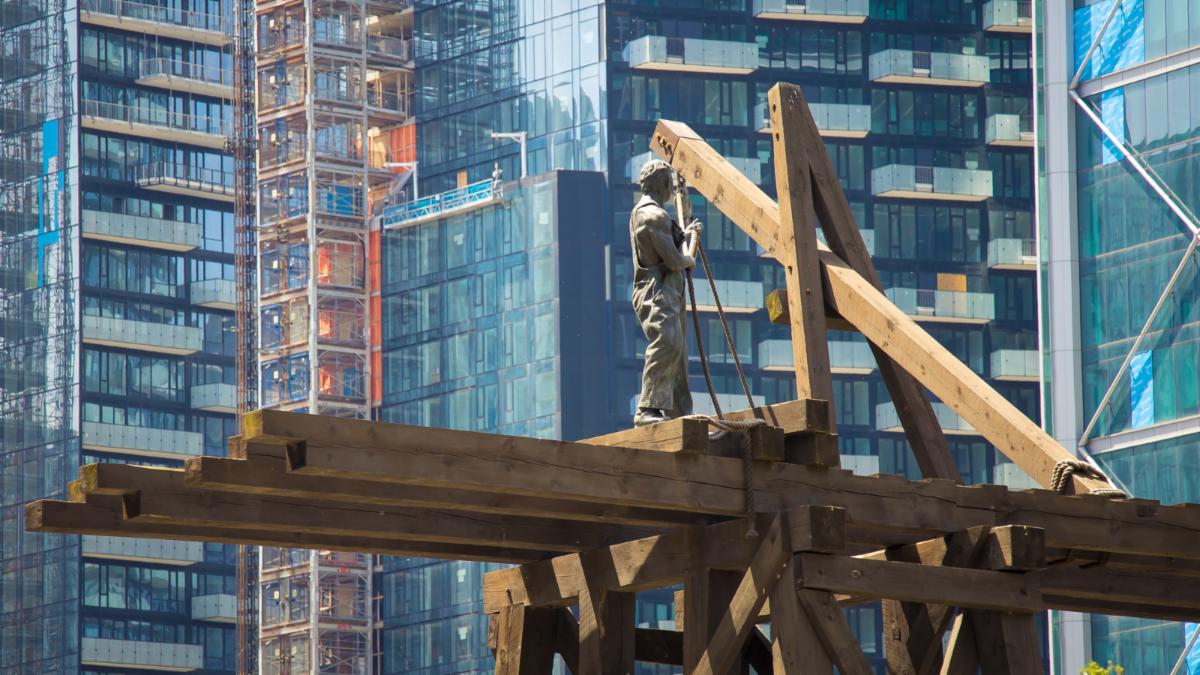 Sculpture of a man working on and positioning large wooden beans with rope. In the background large multi storey buildings are visible.