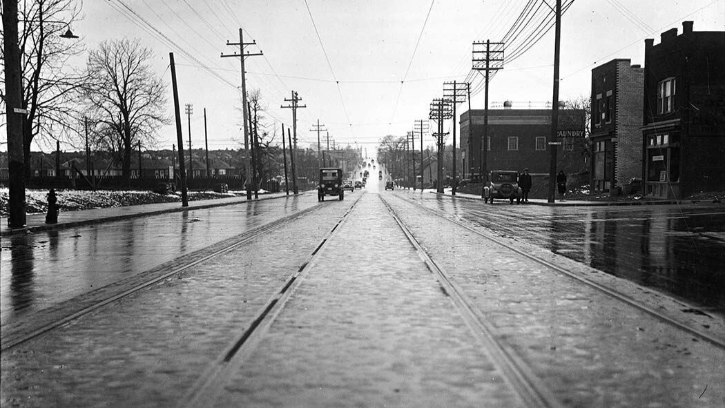 Black and white photograph of street from ground level. The photo is centered around the streetcar tracks with only a few cars on the road in the distance. The ground is wet with a mixture of snow and rain.
