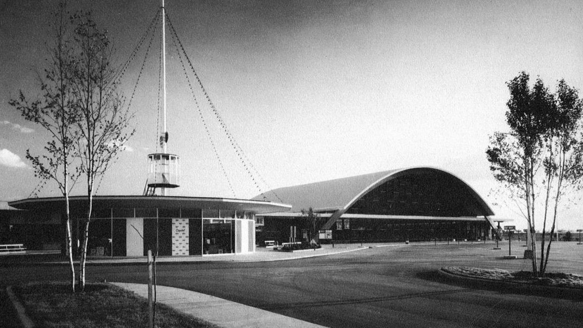 Black and white image of low storey building. To the right a long domed structure appears. To the left a circular pavilion like entrance is visible with a long tower rising from the centre.