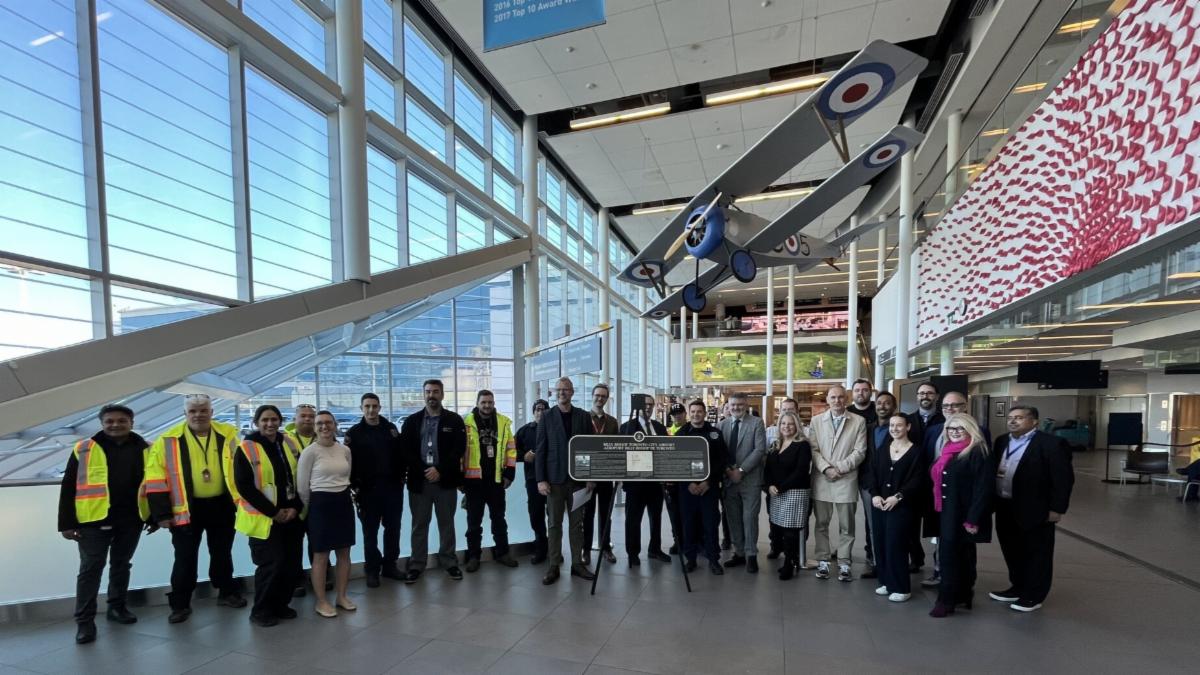 A horizontal line of people stand behind the newly unveiled plaque on an easel posing for a photo.
