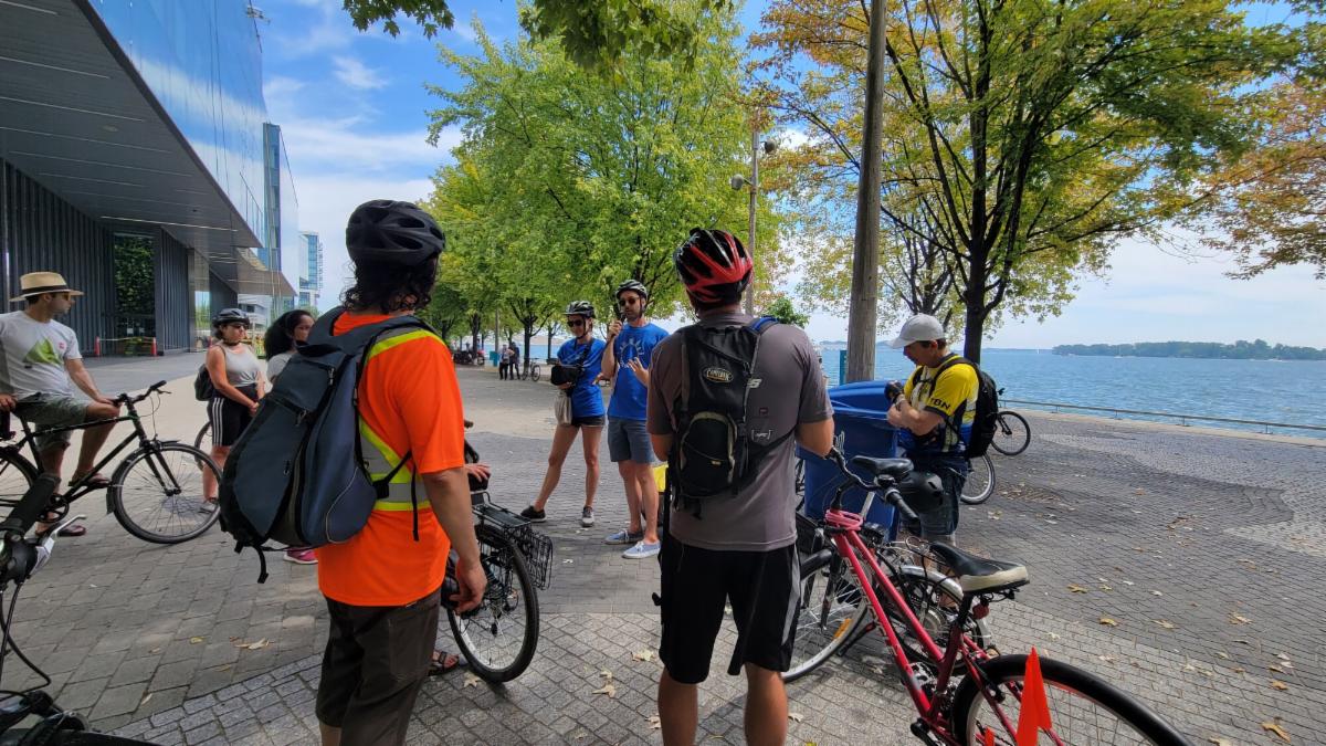 A group of individuals with bikes gather on a concrete sidewalk near a lake. There is a large building in the background along with some green trees.