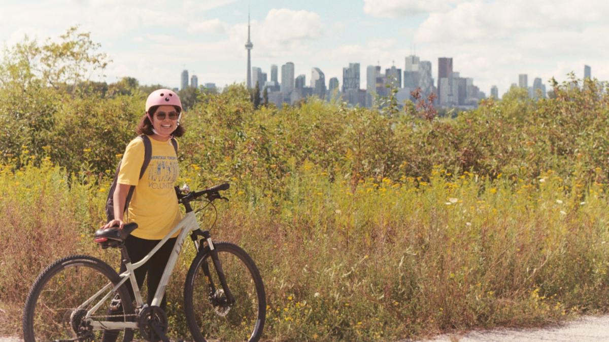 A volunteer wearing a helmet and yellow Heritage Toronto t-shirt stands beside a bicycle. She is facing the camera and smiling. Behind her is an area covered in greenery. In the background of the photo the Toronto city skyline can be seen.
