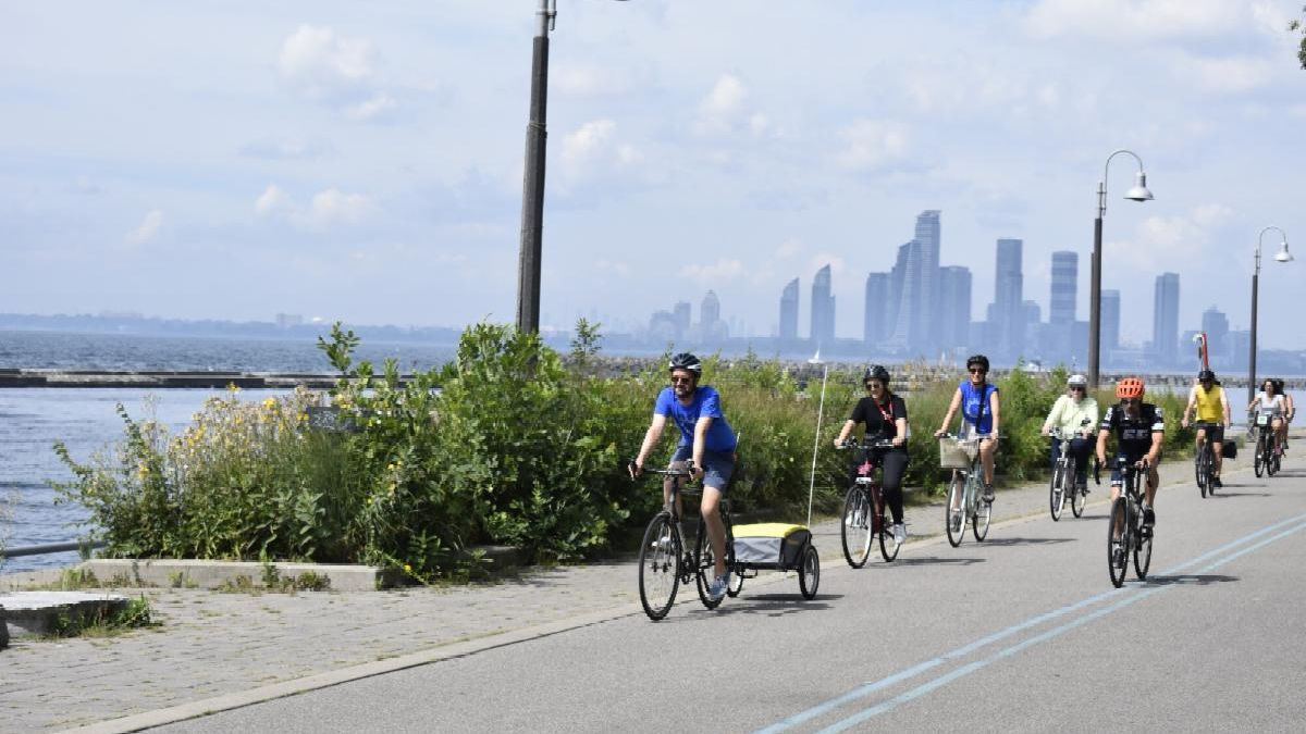 Line of cyclists riding on the side of a round next to a waterfront. In the distance a cityscape is visible of tall skyscrapers.