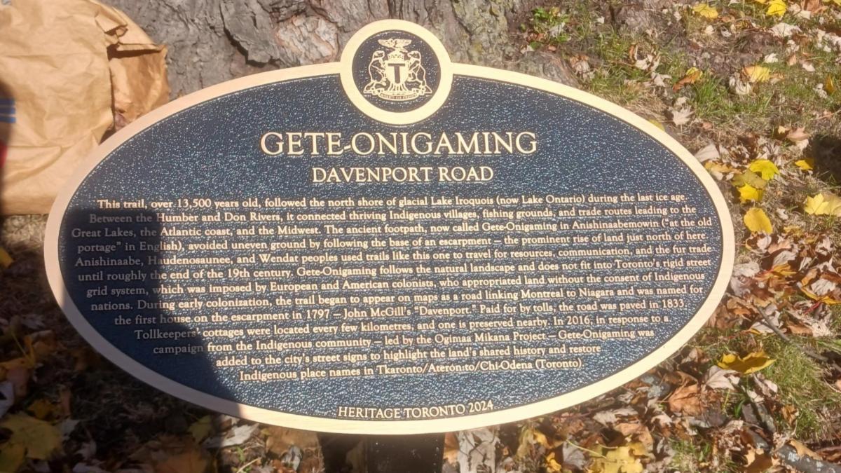 Image of plaque outline commemorating the portage trail known as Gete-Onigaming. There are fallen leaves in the background.