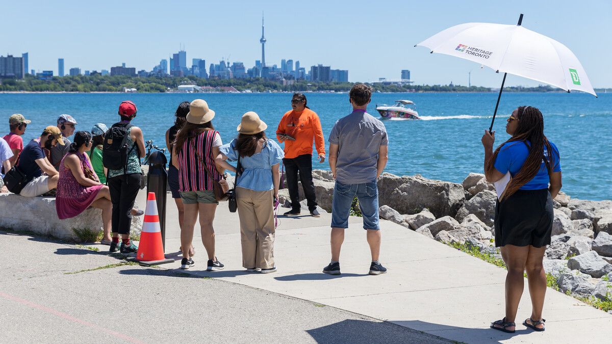 Group of people stand and sit along a path that fronts a waterfront view with a city skyline showing the CN tower behind. A person carrying a white umbrella appears on the right.