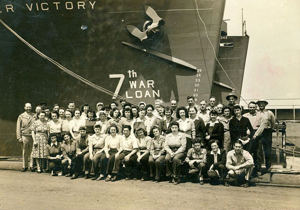 Group of women and men shipyard workers posed in front of a ship displaying a 7th War Loan sign during World War II at a Kaiser Shipyards