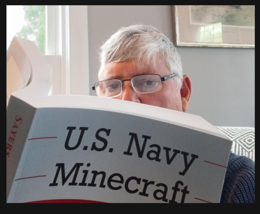 David Winkler, a man with gray hair and glasses, sits indoors reading a book titled "U.S. Navy Minecraft.
