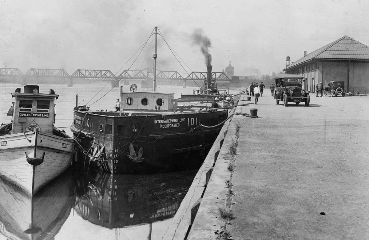 black canal ship ILI tied up alongside a building with tugboat to the right