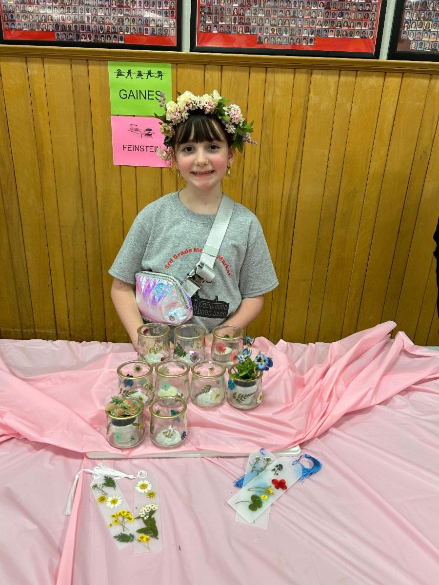 A Mercer third-grader poses wearing a flower headband at a table with her products, candle holders and bookmarks decorated with flowers.