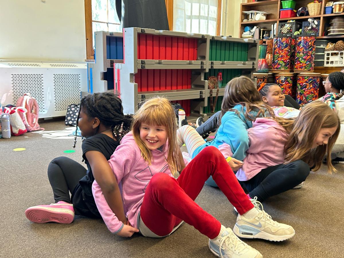 Two students sit on the floor at Lomond with their backs facing each other and lock their arms, trying to work together to stand up at the same time.