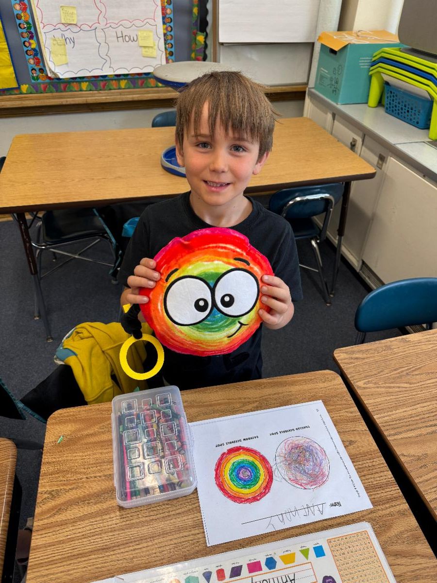 A student sits at a desk holding a rainbow colored toy and smiles.