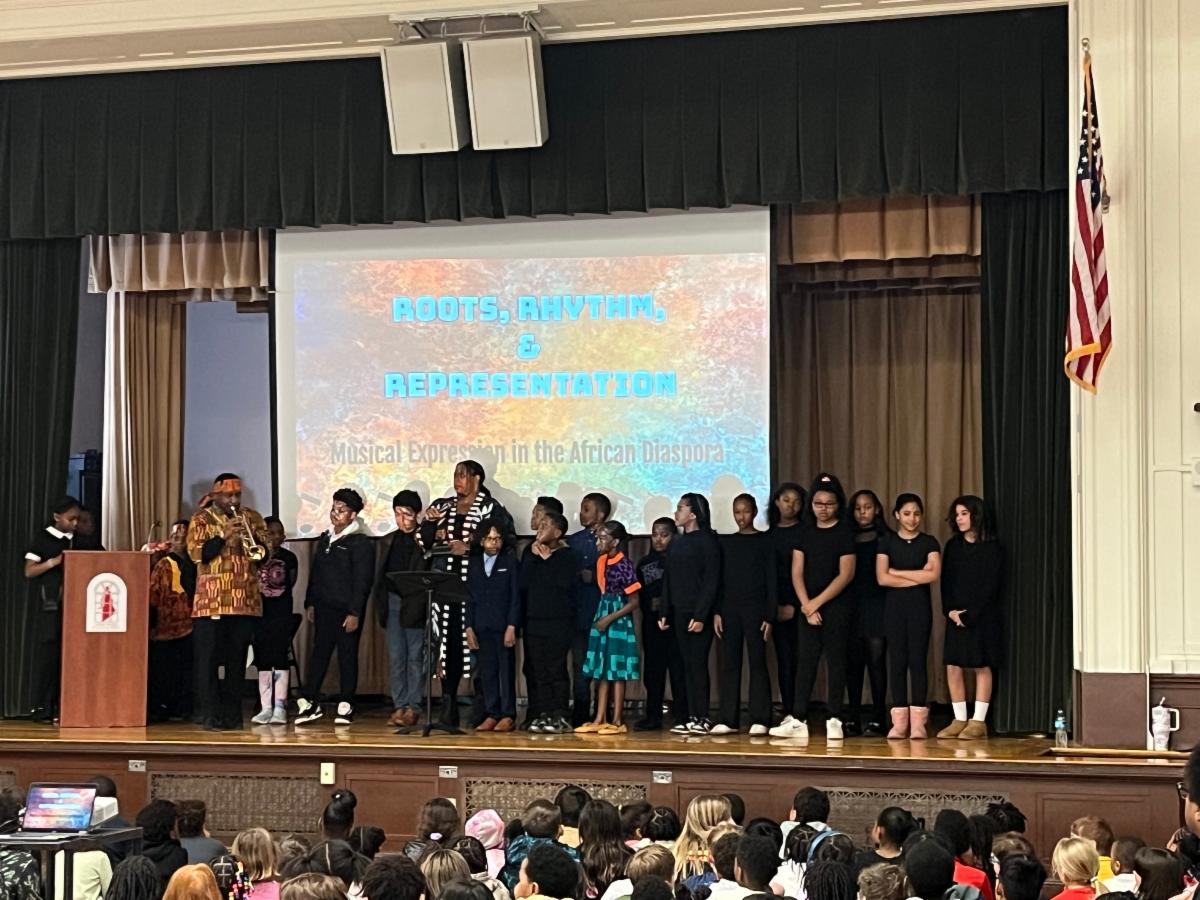 Students sing on stage with Teacher Alesha Edwards and Student Support Coordinator AJ Jennings.