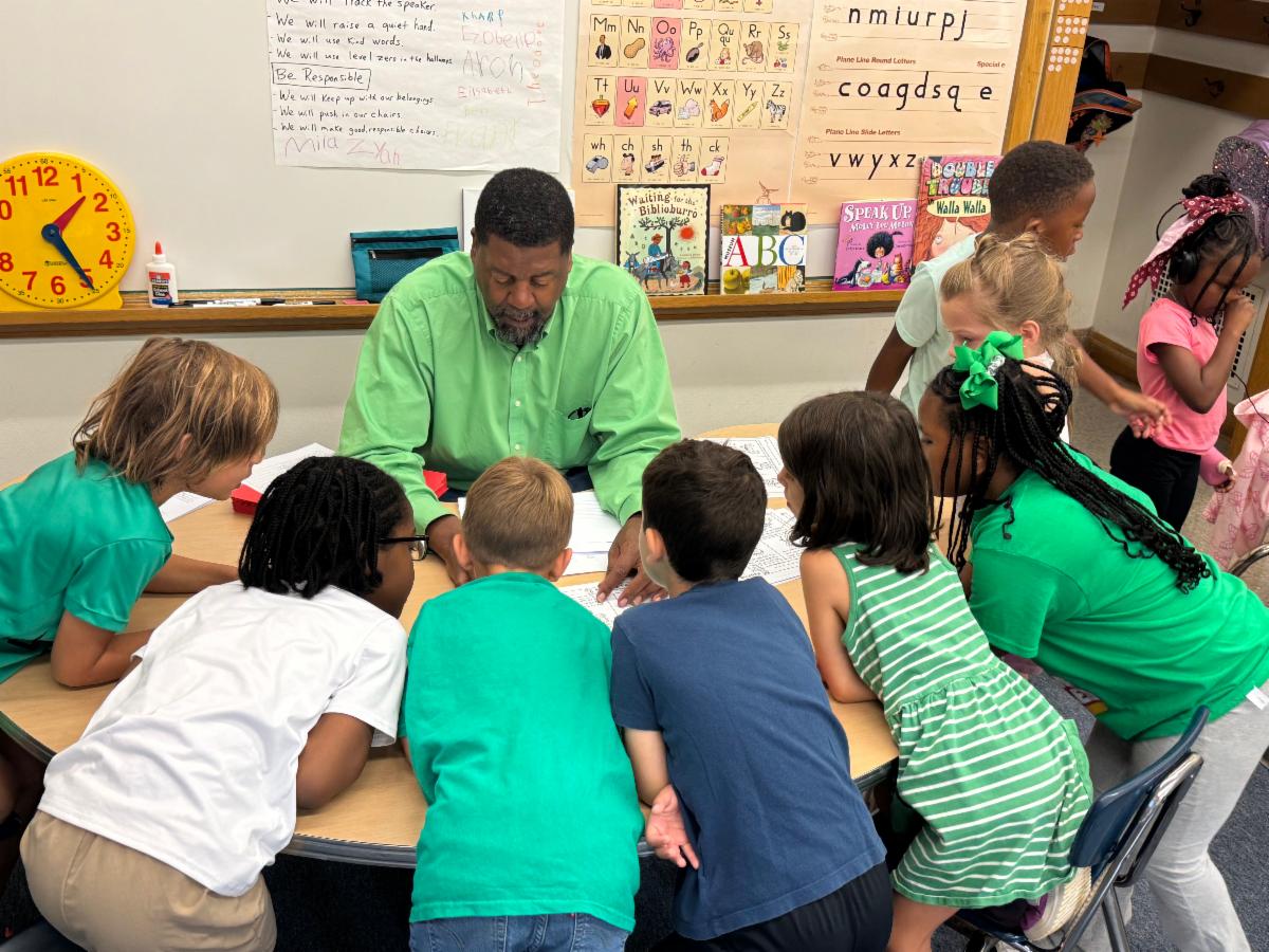 Students dressed in green surround a table while talking to their teacher.