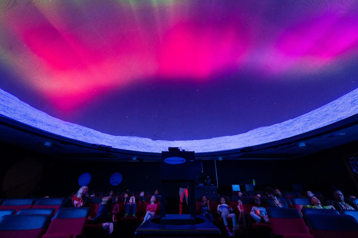 This picture of a planetarium shows a class sitting underneath a pink and purple sky.