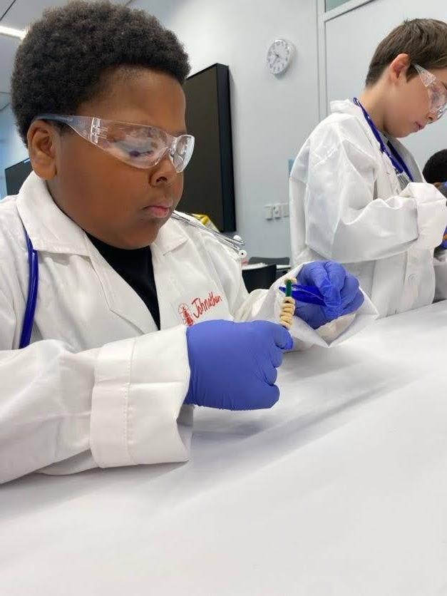 A student sits at a table wearing a white lab coat, plastic gloves, and protective eyewear.
