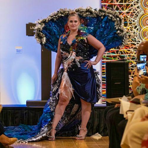 Creating Ocean Legacies display piece. The piece shows a knee-length and shiny blue dress with a train and colorful neckline. The dress has a fanned part behind its model and a 3D fish element in the center of the dress. It is worn with sparkly gray heels.