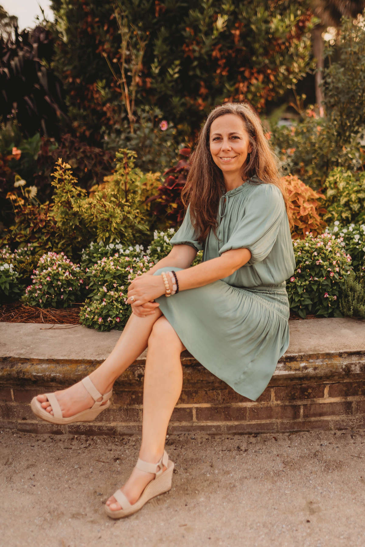 Tina Hartford in a sage green dress sitting on a stone bench with a garden in the background