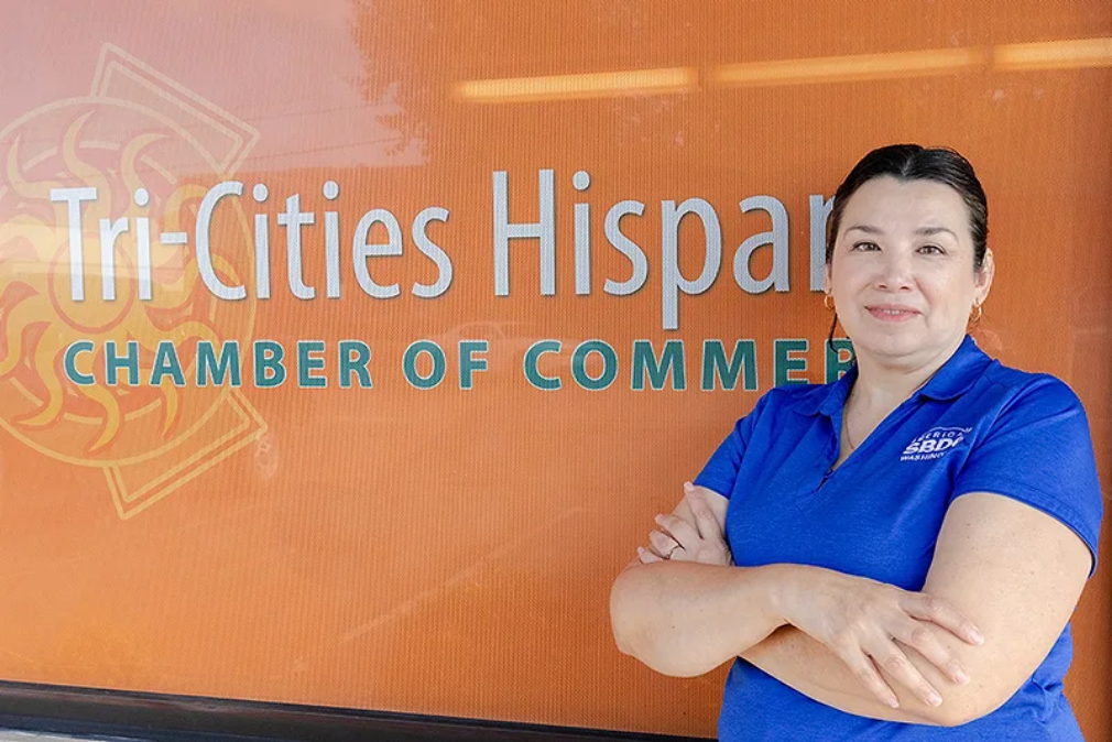 Woman in blue shirt standing in front of Tri-Cities Hispanic Chamber of Commerce logo.