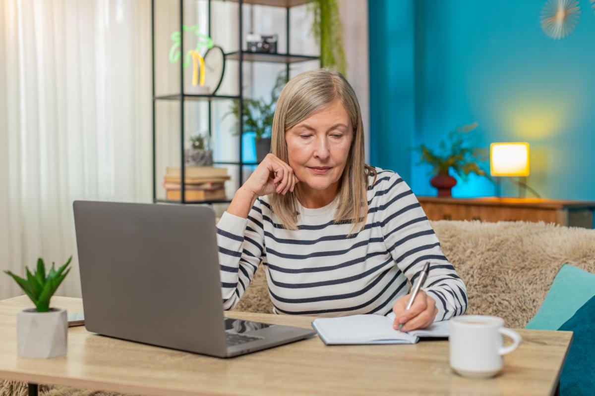 Mature woman participating in online course on laptop. Caucasian grandmother taking notes while sitting on sofa at home table engaging in learning activities enhancing knowledge education lessons
