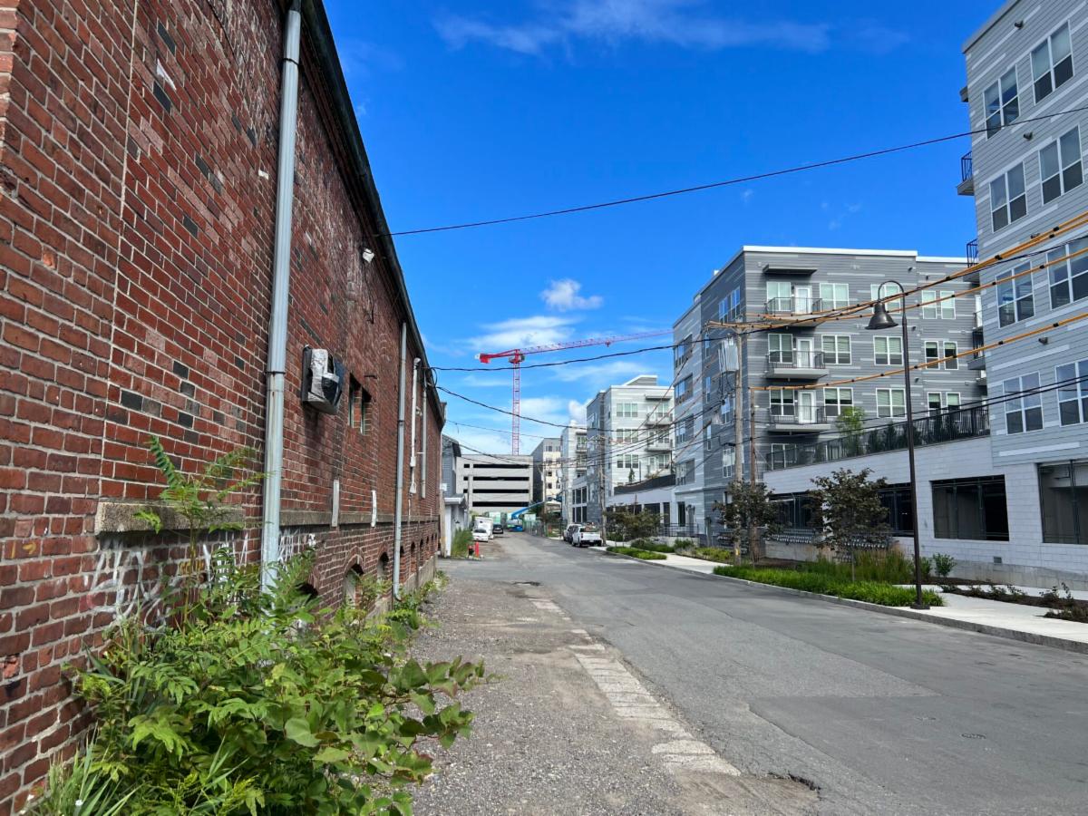 Area that is part of the Chelsea Everett Greenway Connector project. An asphalt road with a large brick building on the left. On the right are newer constructed apartment buildings. The road is leading to a large parking garage.