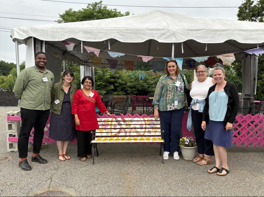 Six people gathered around an elaborately painted bench - three on each side in front of a large white tent.
