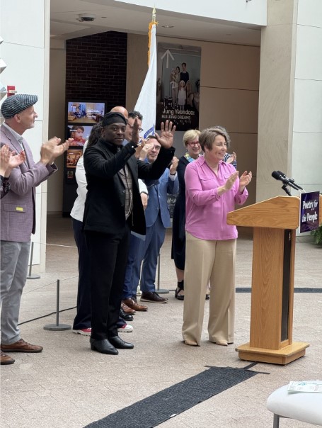 Regie Gibson, wearing a black suit, stands in front of a podium as Governor Maura Healey and others applaud him during the ceremony marking his appointment as Massachusetts’ inaugural Poet Laureate. A state flag is visible in the background. 