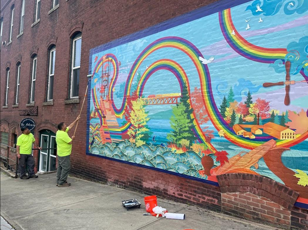 workers in bright green shirts apply finishing touches to a colorful mural on the side of a brick building the mural features rainbows trees water a bridge and small buildings in a vibrant landscape scene