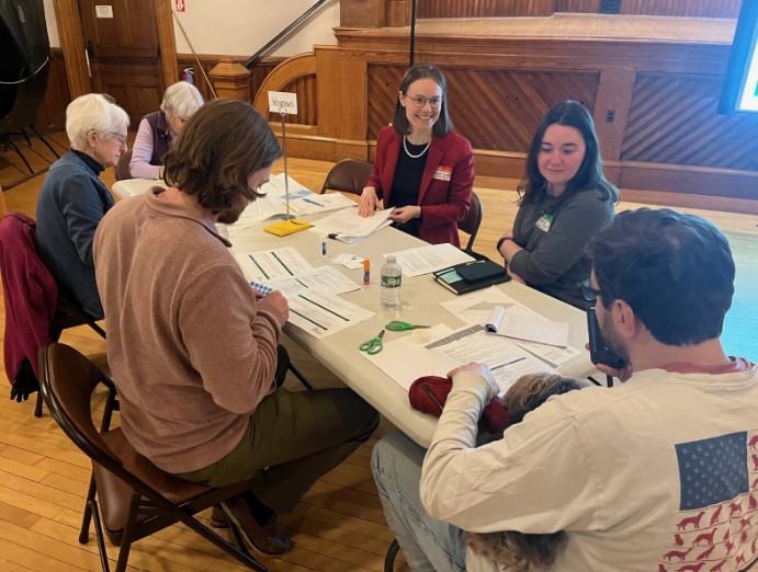 A group of six people is seated around a table in a wood-paneled room, engaged in a discussion. The table is covered with papers, pens, scissors, glue sticks, and other materials. Two women, one wearing a red blazer and another in a gray sweater, are smiling and interacting with the group. One participant holds a small dog in their lap. A small sign on the table reads "Windows," indicating the topic of their discussion. The participants appear focused and engaged.
