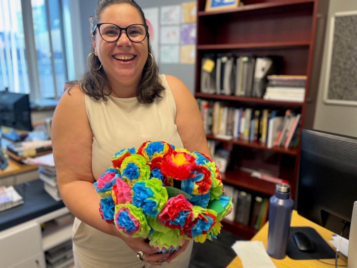 Smiling woman in a beige outfit holds a colorful tissue-paper flower bouquet in an office.