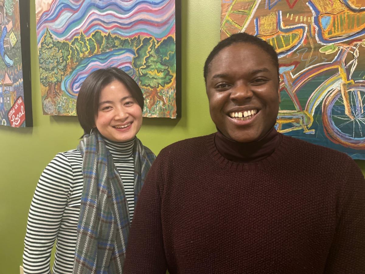 A photo of two smiling individuals standing in front of vibrant, colorful artwork. On the left is Nubes Chen, wearing a striped shirt and plaid scarf. On the right is Lafayette Cruise, wearing a burgundy sweater. 