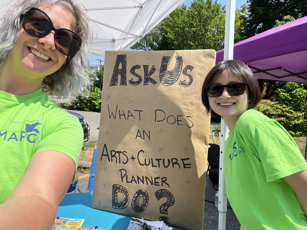 two women in bright green shirts smile at the camera while standing beside a handmade sign that reads Ask Us What Does an Arts and Culture Planner Do at the Arts and Culture Teams table during the Back to School Extravaganza in Framingham