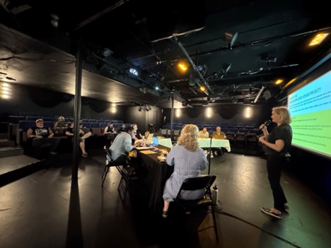 A woman with a microphone standing in front of a screen - presenting to a group of seated people in a room painted black.