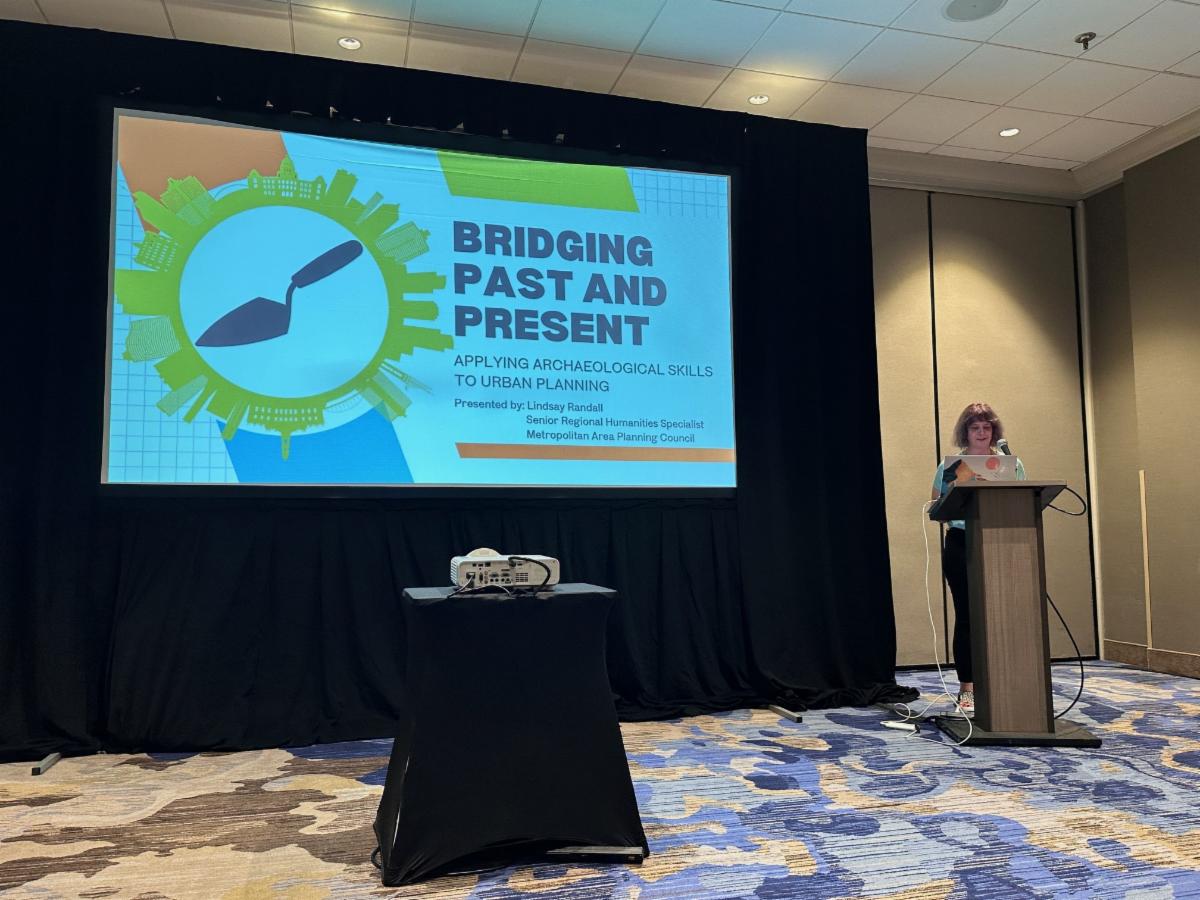 A woman stands at a podium giving a presentation in a conference room. Behind her is a large projector screen displaying a colorful slide titled "Bridging Past and Present: Applying Archaeological Skills to Urban Planning."    