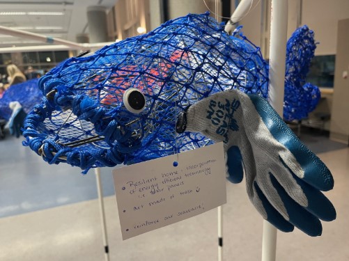 A blue wire mesh fish sculpture with googly eyes and a work glove attached to its side, labeled "Save Our Shores." A handwritten card hangs nearby, sharing reflections on resilient home design, climate preparedness, and seasonal awareness. 